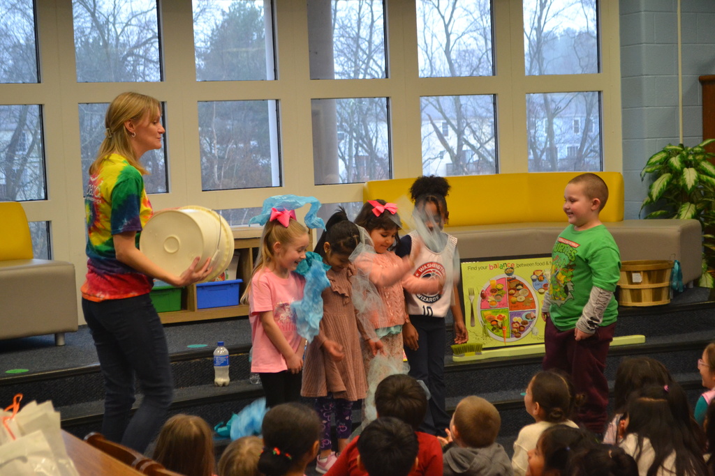Students laugh as a presenter holds a large white bucket and blue fabric props during a hands-on activity about dental hygiene. A young boy stands facing the group while classmates observe.