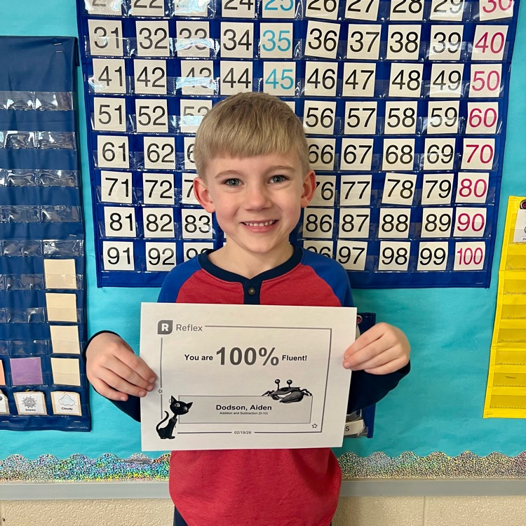 a blonde haired first grade boy with a blue and red shirt smiles proudly in front of a 100 chart holding a certificate that reads “100% fact fluent”  