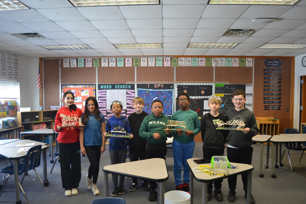 Eight elementary school students stand in a classroom holding handmade bridge models constructed from craft sticks and glue. The classroom walls display learning posters and a word search board. Desks are arranged around the room, and the students are posing together in front of their projects.