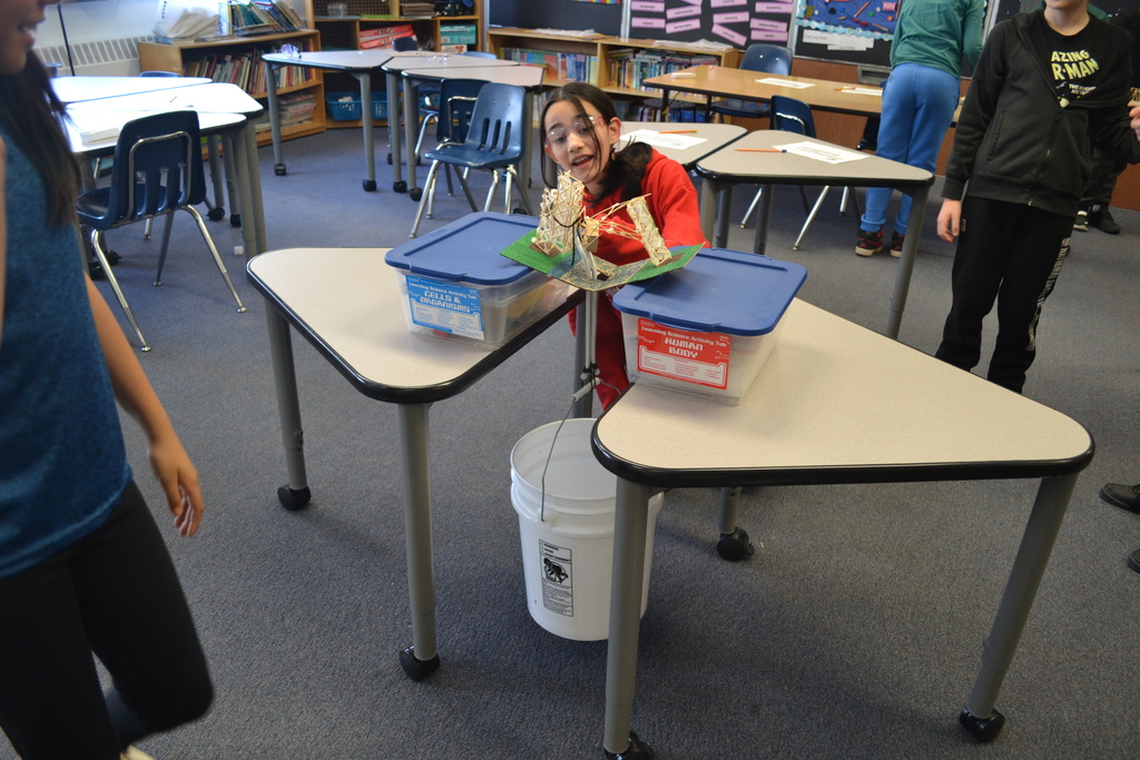 A student wearing safety goggles leans forward to observe a bridge model placed across two plastic storage bins on desks. A string extends from the center of the bridge to a white bucket on the floor, indicating a load test. Other students stand nearby watching, and classroom desks and posters are visible in the background.