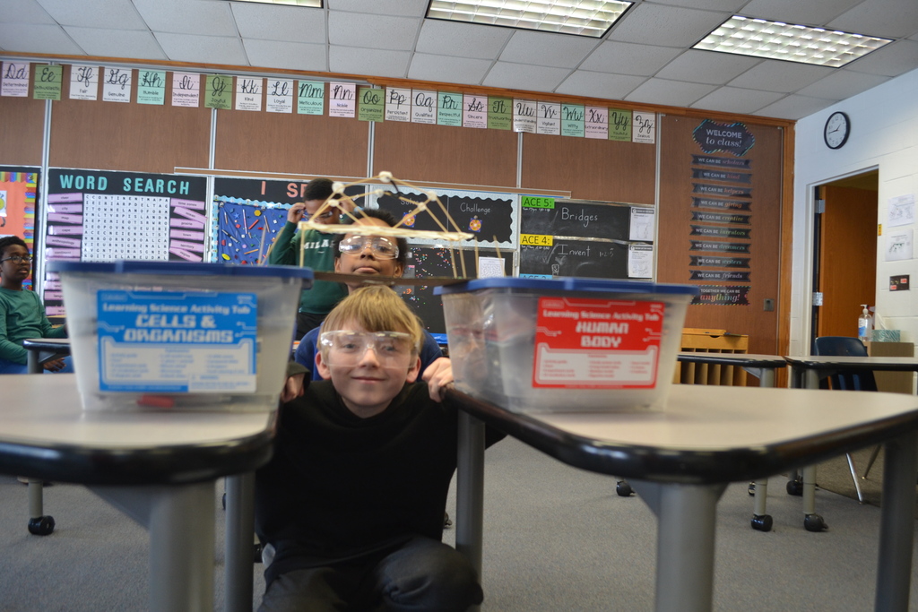 A student kneels between two desks while another student stands behind, as a bridge model rests across plastic bins placed on top of desks. The students are wearing safety goggles, and the classroom bulletin boards and posters are visible in the background.