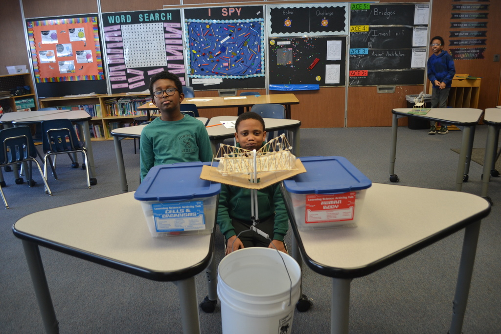 Two students sit behind a handmade bridge model positioned across two plastic storage bins placed on separate desks. A string hangs from the center of the bridge to a white bucket on the floor, likely for weight testing. Classroom desks, chairs, and bulletin boards are visible in the background, with another student standing near the back of the room.
