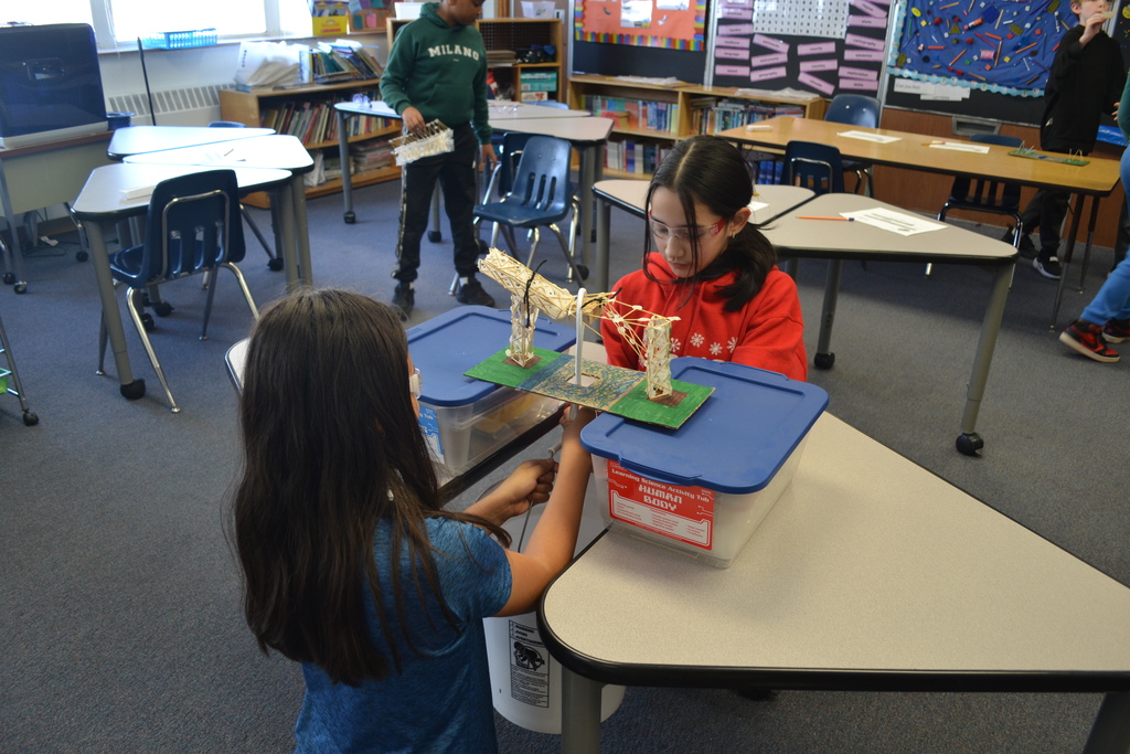 Two students work together at a desk on a bridge model supported by plastic storage bins. One student steadies the structure while the other adjusts part of the model. Additional students and desks are visible in the classroom behind them.