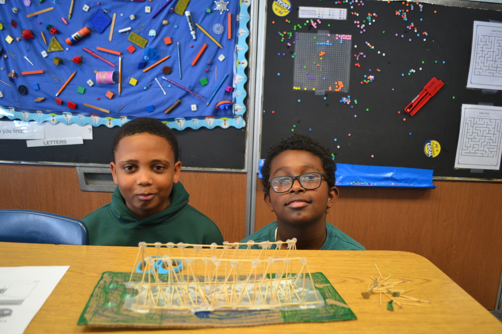 Two students sit at a desk with a completed bridge model displayed in front of them on a green base. Small craft materials lie beside the structure. Classroom bulletin boards with colorful displays are visible in the background.
