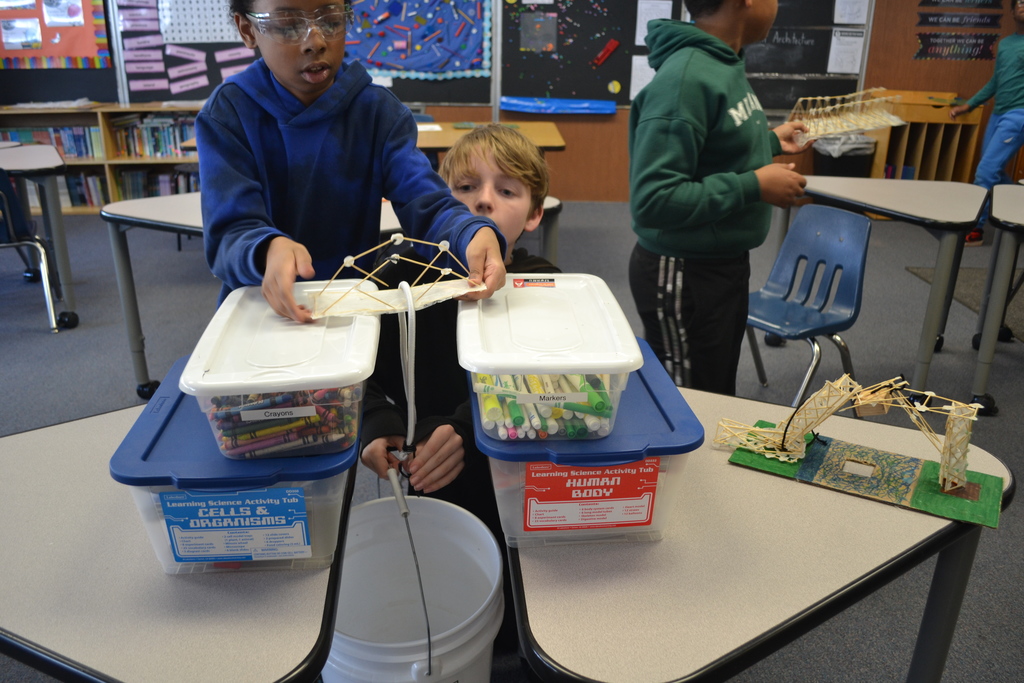 A student wearing safety goggles places a handmade bridge model across two plastic storage bins positioned on separate desks. The bins are labeled “Cells & Organisms” and “Human Body.” A string hangs from the center of the bridge down to a white bucket on the floor for weight testing. Another student kneels behind the setup, watching closely, while additional students and completed bridge models are visible in the classroom.
