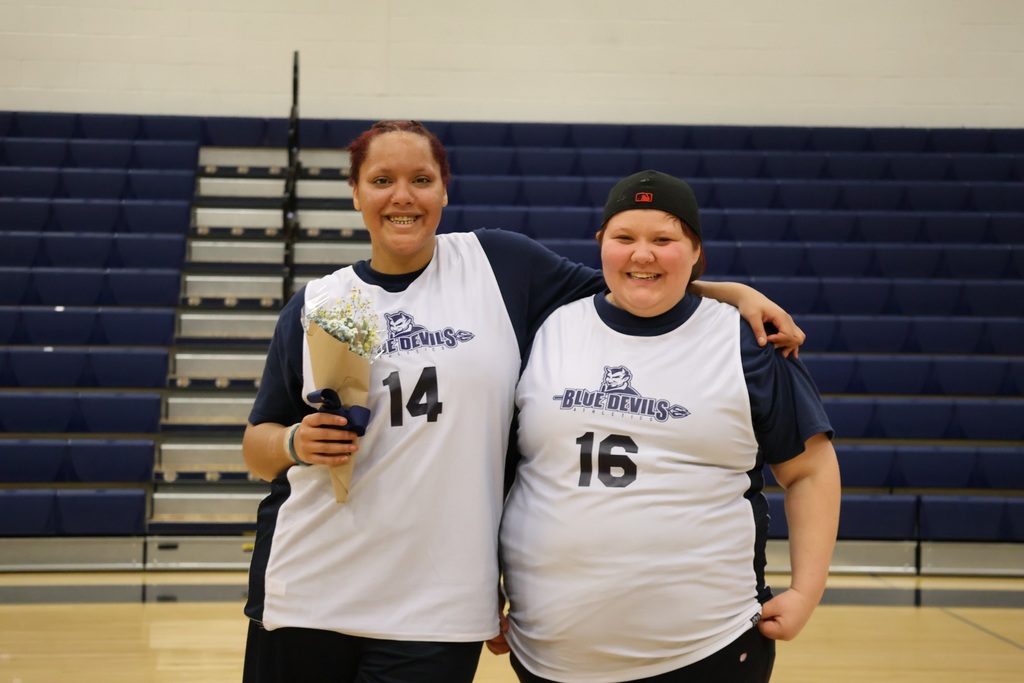 Two students in blue devils basketball jerseys pose side by side for a picture. One is wearing a hat and the other holds flowers.