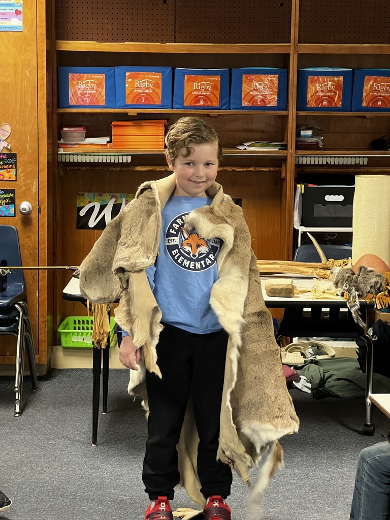A student stands smiling while wearing an animal pelt draped over their shoulders. A classroom shelf with labeled book bins and additional cultural artifacts is visible behind them.