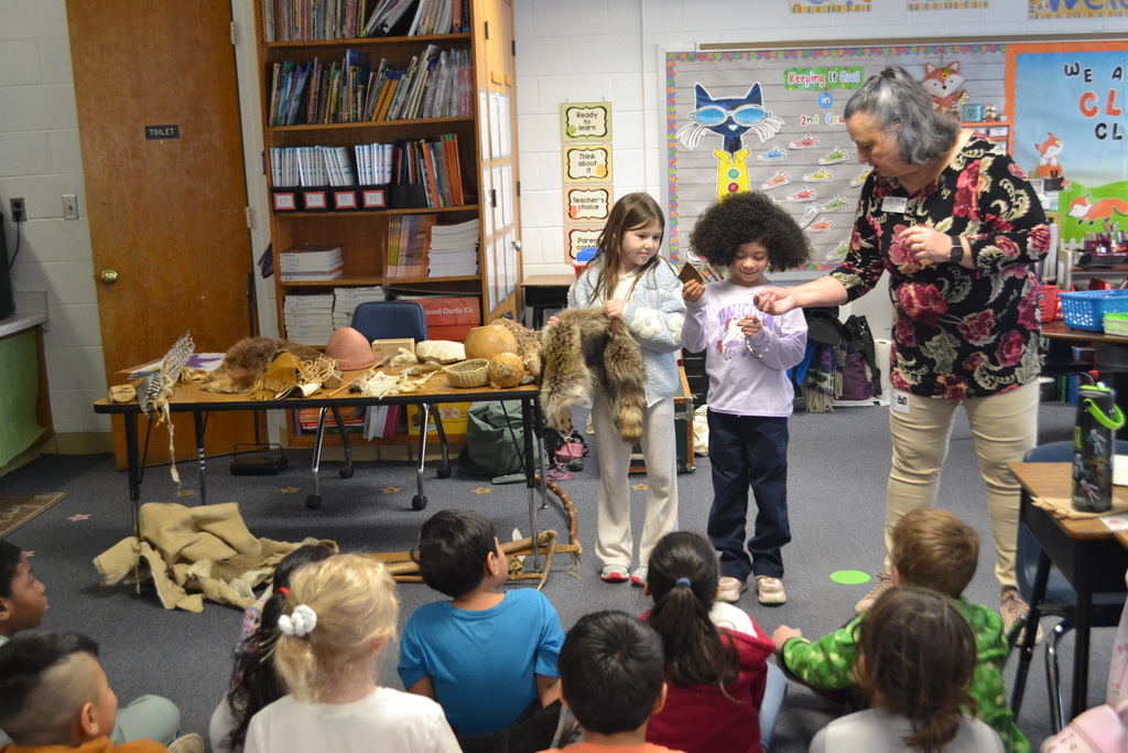 Two students stand at the front of the classroom holding animal pelts while a guest presenter stands beside them speaking to seated classmates. A display table behind them holds additional pelts, gourds, baskets, and tools. Students sit on the floor listening.