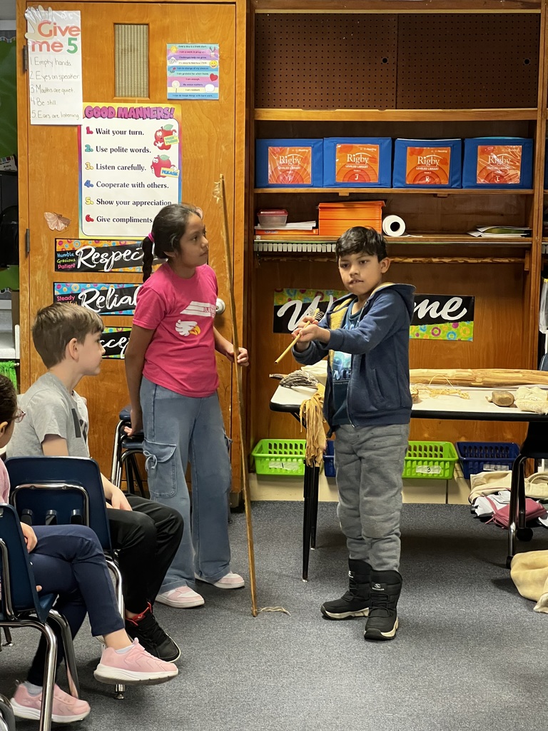A student stands at the front of the classroom holding a long wooden tool while classmates sit nearby watching. A classroom door behind them displays posters about good manners and classroom expectations.