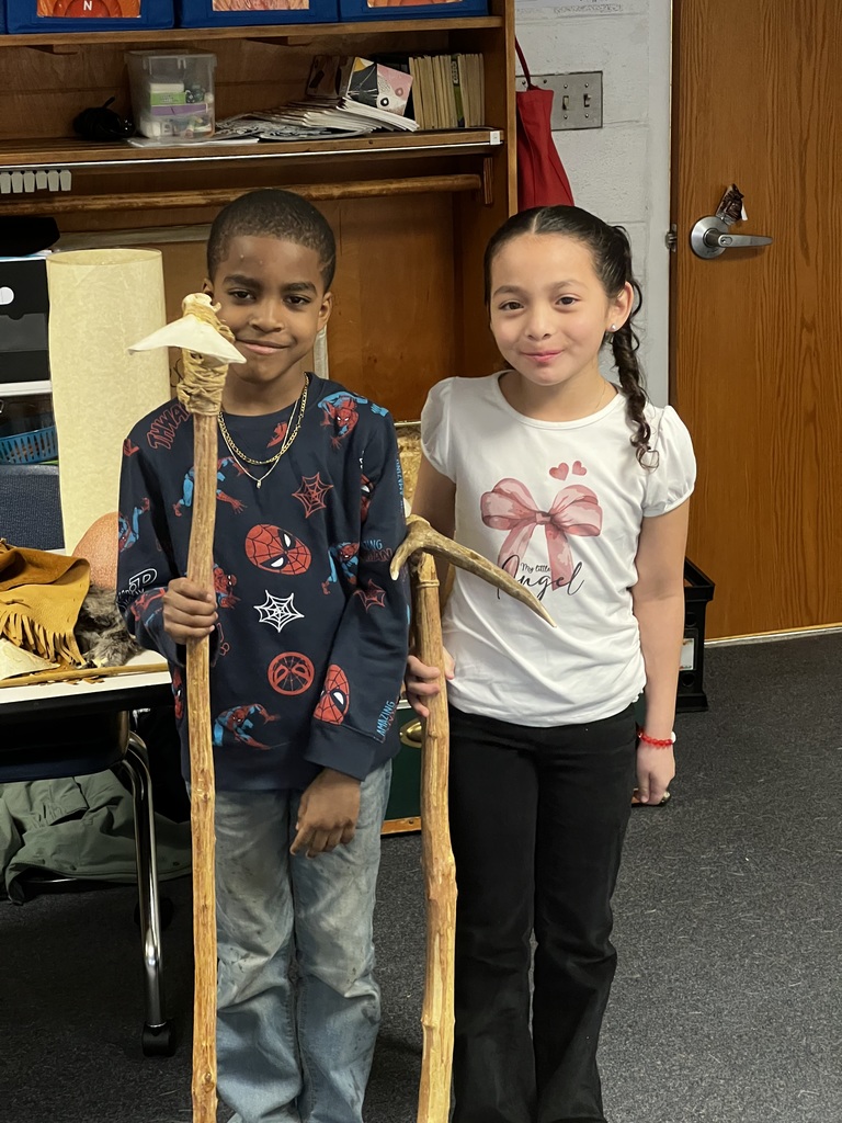 Two students stand side by side holding long wooden tools and smiling at the camera. A classroom shelf with labeled book bins and cultural artifacts is visible in the background.