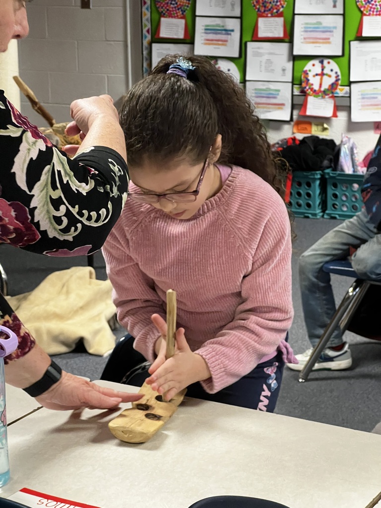 A student sits at a desk using a wooden tool to press into a small wooden board while an adult guides their hands. Classroom artwork and supplies are visible in the background.