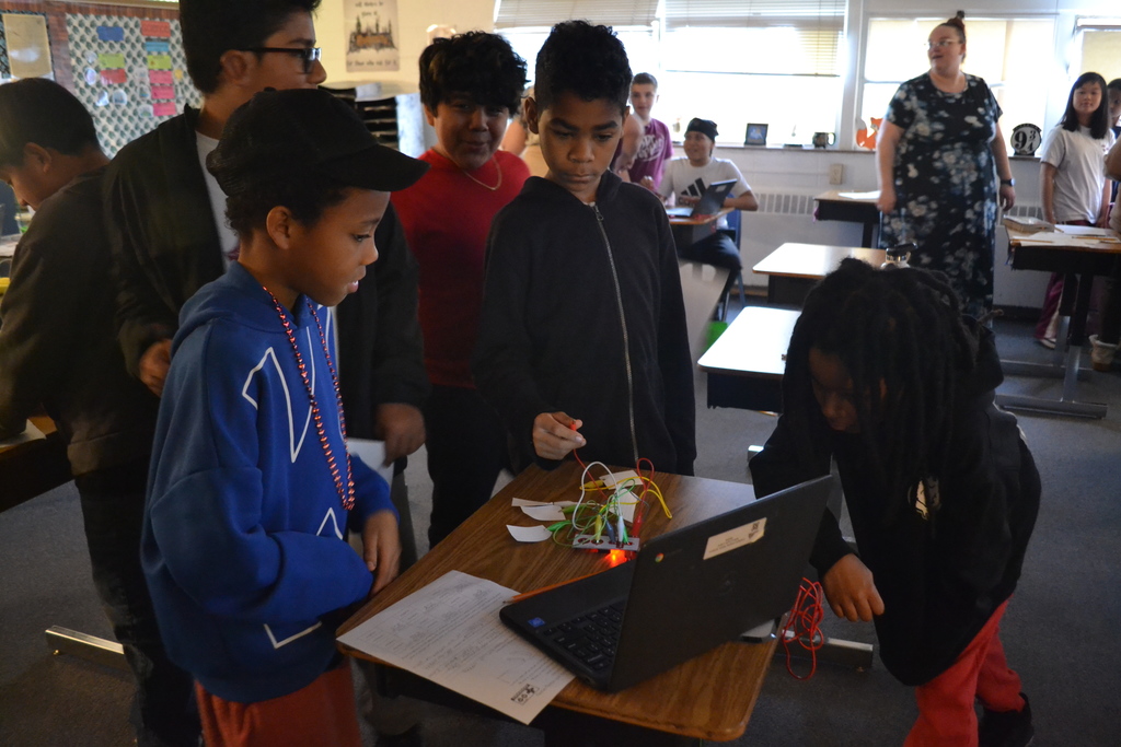 Several students gather around a desk with a laptop and a small electronic circuit board connected by colorful wires. One student holds a wire while others watch closely. The teacher stands in the background near the windows.