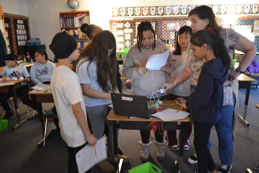 A teacher stands with a group of students gathered around a desk with a laptop and electronic components connected by wires. Students hold worksheets and pencils while discussing the activity together.