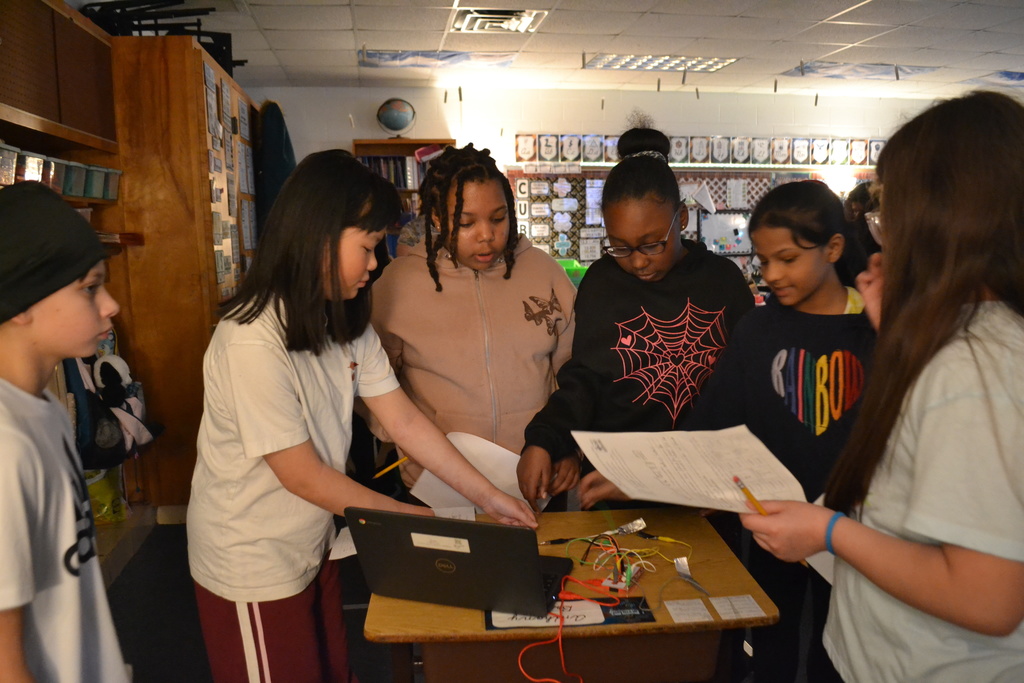 A group of students stand closely around a desk reviewing papers while working with a laptop and a wired circuit board. They appear to be collaborating on a classroom technology project.