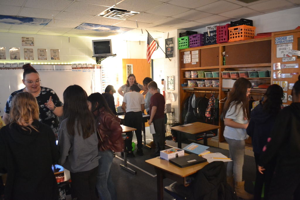 Students move around the classroom working in small groups at desks. A teacher stands to the left speaking with students while others gather near the back of the room. Classroom shelves, posters, and an American flag are visible.