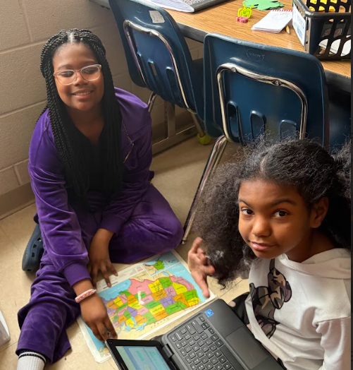 Two students sit on the floor next to desks with a United States map spread out in front of them. One student points toward a laptop screen while the other looks up at the camera. A small robot sits on the map.