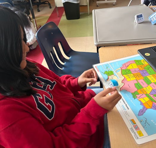 A student in a red sweatshirt sits at a desk holding a small round robot above a United States map. A laptop rests nearby on the desk.
