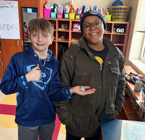Two students stand side by side in a classroom smiling at the camera. One student holds a small round robot in their hand. Classroom shelves and supplies are visible behind them.