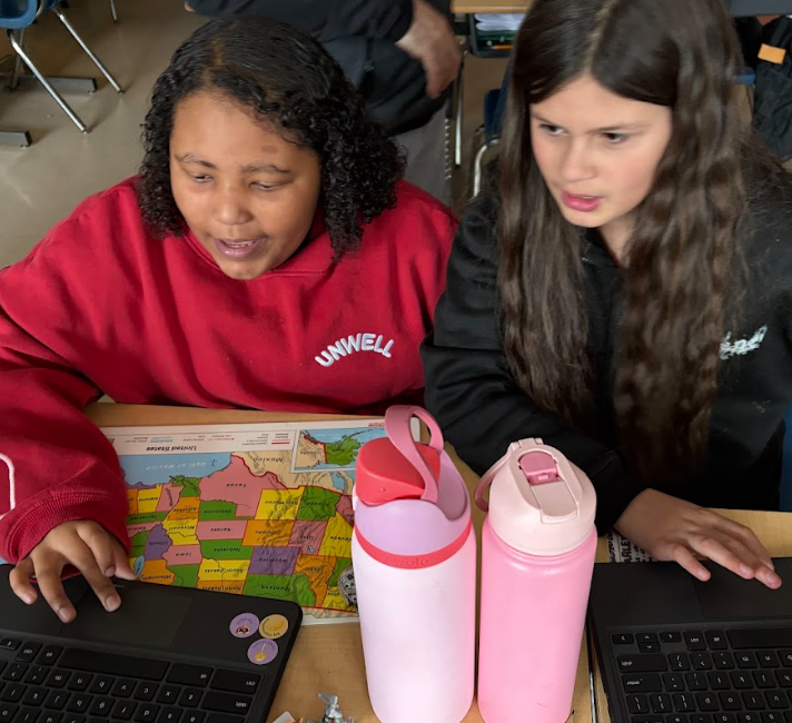 Two students sit at a desk working on laptops with a United States map placed between them. Two pink water bottles sit in the foreground. Both students appear focused on their screens.