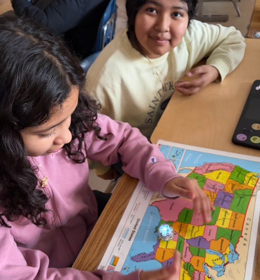 Two students sit at a desk with a United States map and a laptop open in front of them. One student moves a small glowing robot across the map while the other looks on.