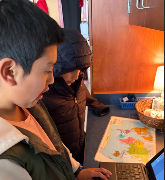 Two students stand at a classroom counter using a laptop next to a colorful world map. One student types while the other looks on. A small glowing robot sits on the map. Cabinets and a lamp are visible in the background.