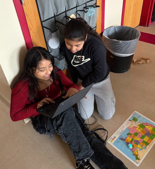 Two students sit on the floor in a hallway using a laptop. A United States map and a small glowing robot are placed on the floor beside them.