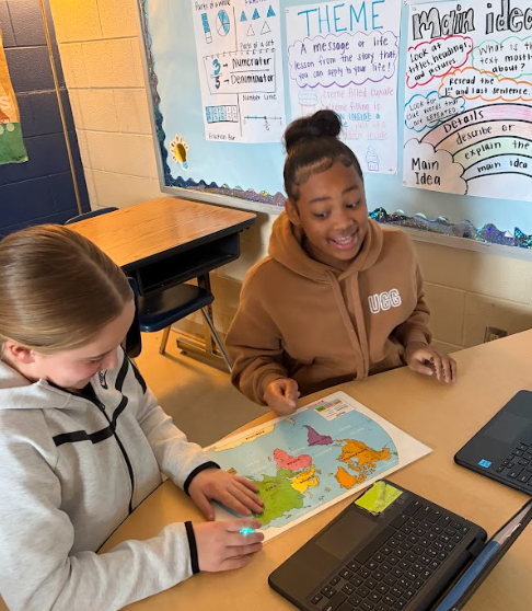 Two students sit at a desk with laptops and a world map. One student moves a small glowing robot across the map while both students smile. Classroom posters about theme and main idea are visible on the wall behind them.