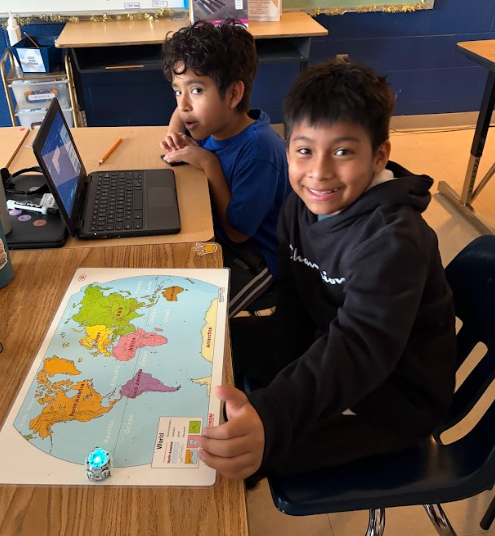 Two students sit at a desk with laptops open and a world map in front of them. One student moves a small glowing robot across the map while the other smiles and watches.