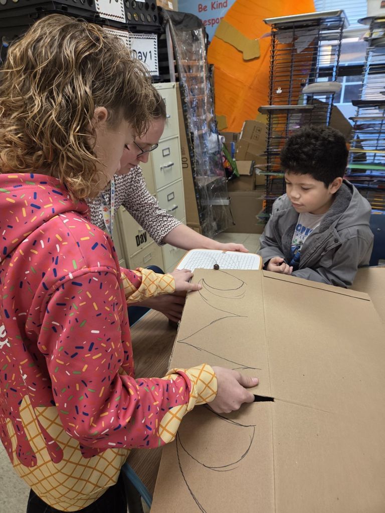 An adult teacher wearing glasses and a patterned shirt assists two students as they maneuver a large, flat cardboard box onto the cutting tool. The child in the sprinkle hoodie holds one side while the teacher helps stabilize the machine.