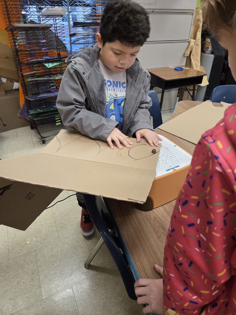 The boy in the grey jacket uses both hands to pivot a large section of cardboard on the white grid surface of the orange cutter, following curved lines drawn in black marker.