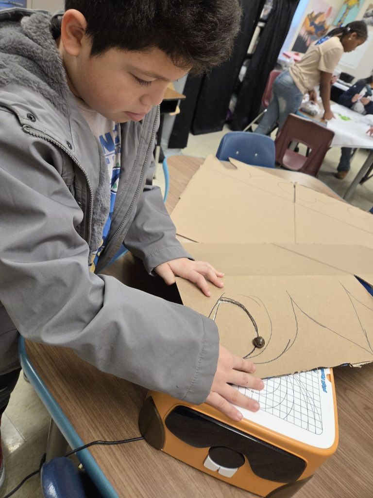 A young boy in a grey jacket over a Sonic the Hedgehog t-shirt focuses intently as he presses down on a cardboard sheet, guiding a circular drawn line through the blade of the orange electric cutting machine.
