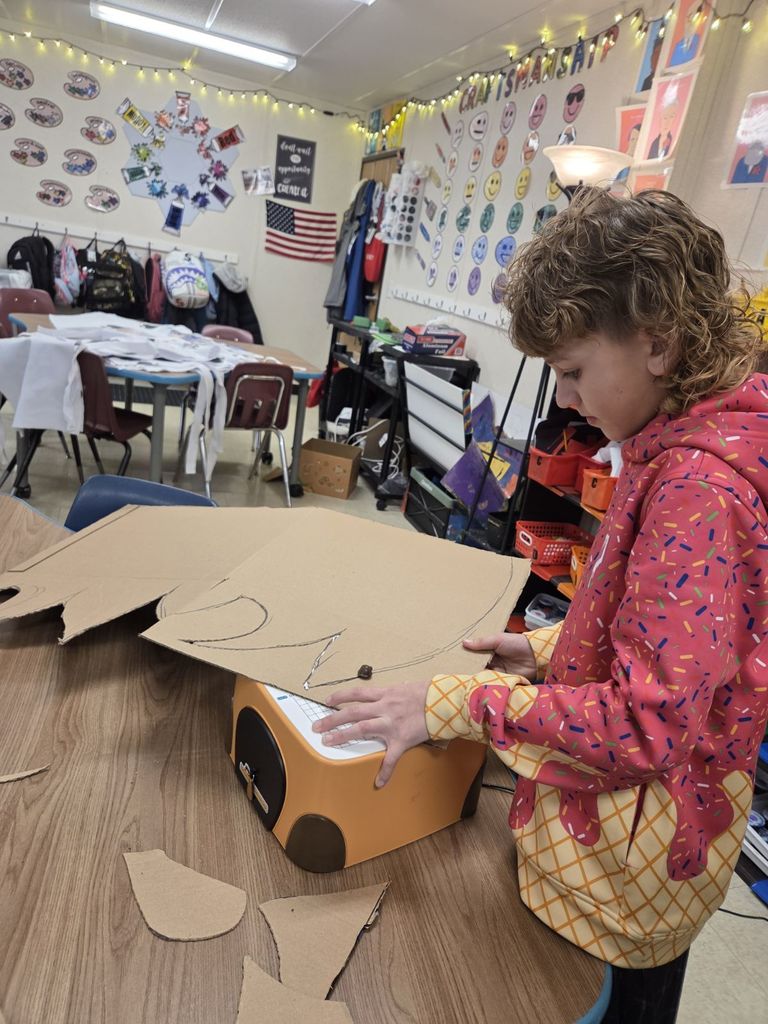 A young child with curly hair and a pink sprinkle-patterned hoodie stands at a classroom table, carefully guiding a large piece of cardboard through an orange electric cardboard cutter. In the background, a colorful art classroom is visible with posters, an American flag, and a string of lights.