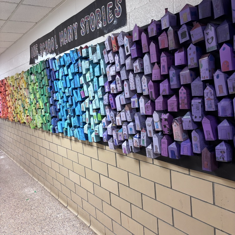 A long school hallway features a large, 3D collaborative art installation titled "ONE SCHOOL MANY STORIES." Hundreds of small, hand-decorated paper houses are mounted on a black background, organized in a vibrant rainbow gradient from green and blue to deep purple.