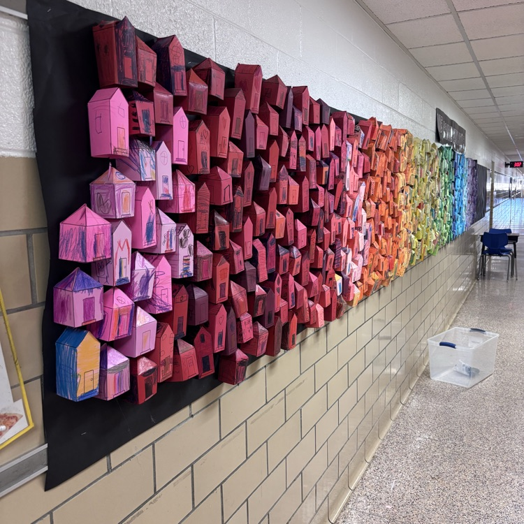 A different angle of the same installation, showing the warm side of the color spectrum. The wall is covered in hundreds of paper houses in shades of pink, red, orange, and yellow, each uniquely colored and drawn on by students.