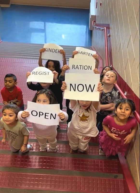 kindergarten children on maroon stairs holding signs on pieces of paper that say kindergarten registration now open