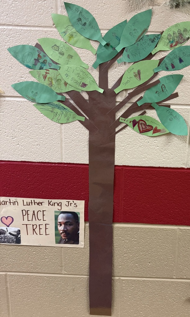 A wall display titled “Martin Luther King Jr.’s Peace Tree” features a large brown paper tree trunk with green paper leaves. Each leaf contains student drawings and messages about love, friendship, peace, and kindness. A small poster with a photo of Dr. Martin Luther King Jr. is placed near the base of the tree on a beige and red brick wall.