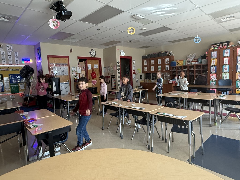 Several elementary students stand beside their desks in a classroom, appearing to participate in an activity. The room has individual desks spaced apart, cabinets and cubbies along the back wall, a clock above the door, and colorful decorations including a birthday board and classroom posters.