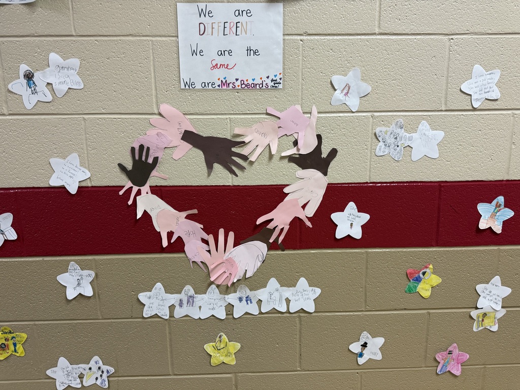 A classroom wall display with a sign that reads, “We are DIFFERENT. We are the same. We are Mrs. Beard’s class!” Below the sign, cut-out paper handprints in various skin tones form a heart shape. Around the heart, student-made star-shaped papers with drawings and short written messages are taped to a beige and red brick wall.