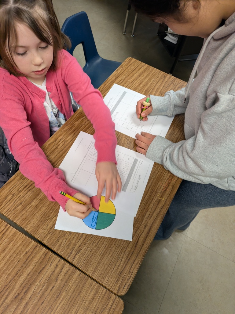 A student in a pink sweater spins a colorful four-section spinner while a partner records tally marks on a probability worksheet during a classroom math activity.