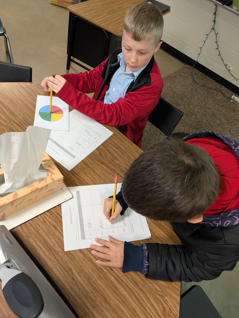Two students work side-by-side at a desk. One spins a four-color paper spinner while the other records tally marks on a worksheet labeled “Spinning Colors.”