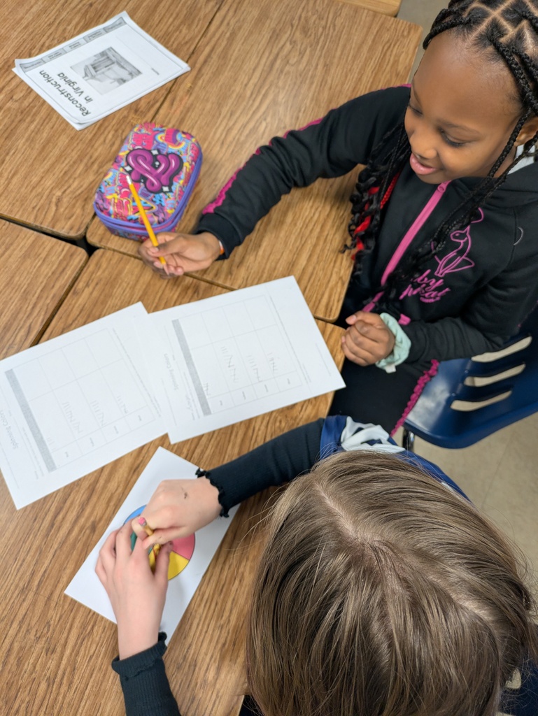 Two students sit at a desk working together on a probability activity. One student holds a pencil above a tally chart while a colorful four-section spinner rests on the desk between them.