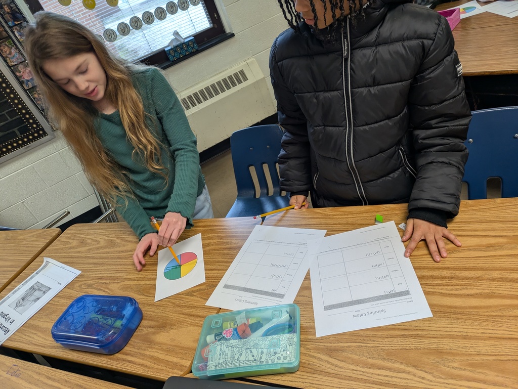 Two students conduct a probability experiment using a four-color spinner and record their results on worksheets. Pencil boxes and classroom materials are visible on the desks around them.