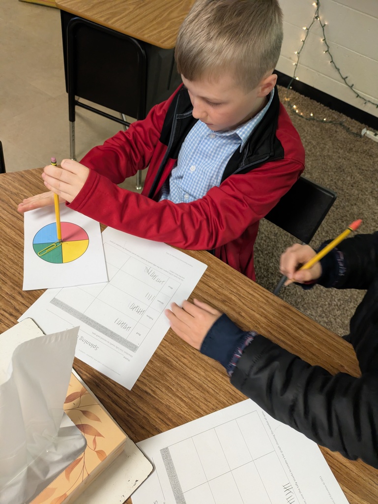 A fourth grade student in a red jacket spins a paper clip spinner divided into four colored sections (red, blue, green, and yellow) while recording tally marks on a probability worksheet at a classroom desk.