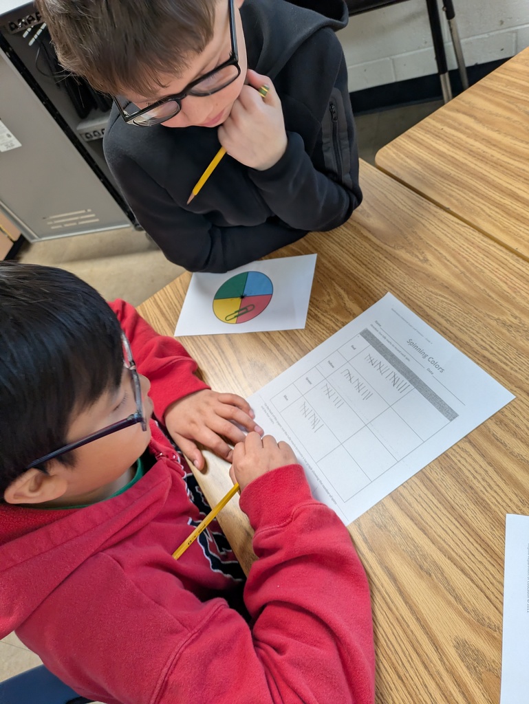 Two students review their results on a “Spinning Colors” worksheet filled with tally marks. A four-color spinner sits on the desk as they discuss their data.