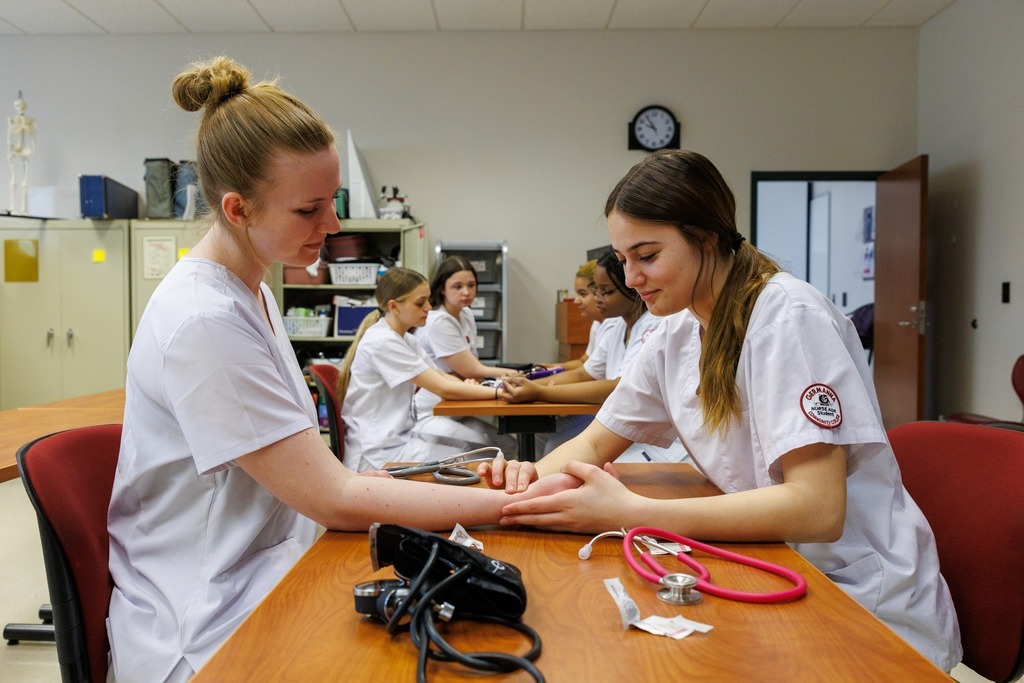 CTEC healthcare technician students in white scrubs practice taking blood pressure and checking vital signs during a hands-on classroom lab.