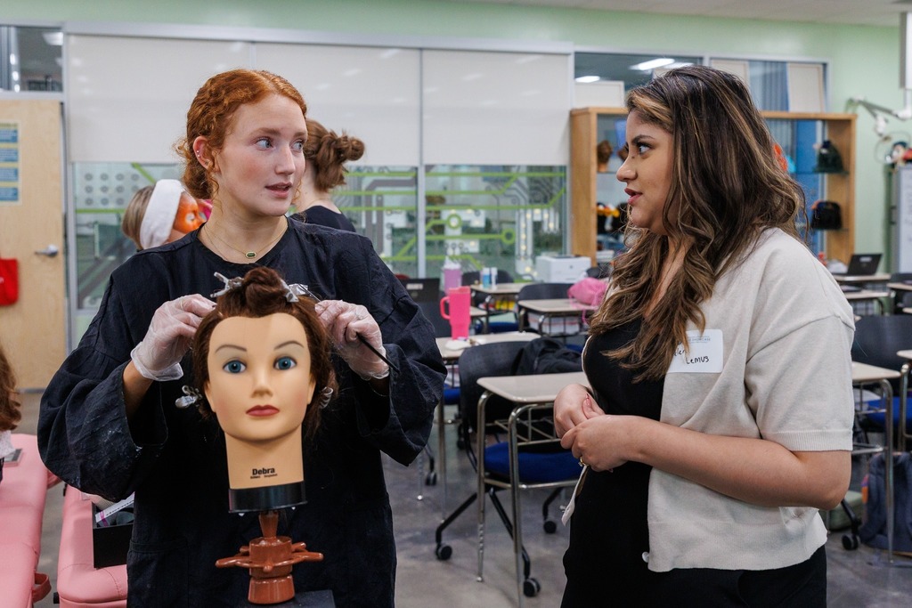 CTEC cosmetology student demonstrates hairstyling techniques on a mannequin while speaking with an industry partner during Skills Showcase.