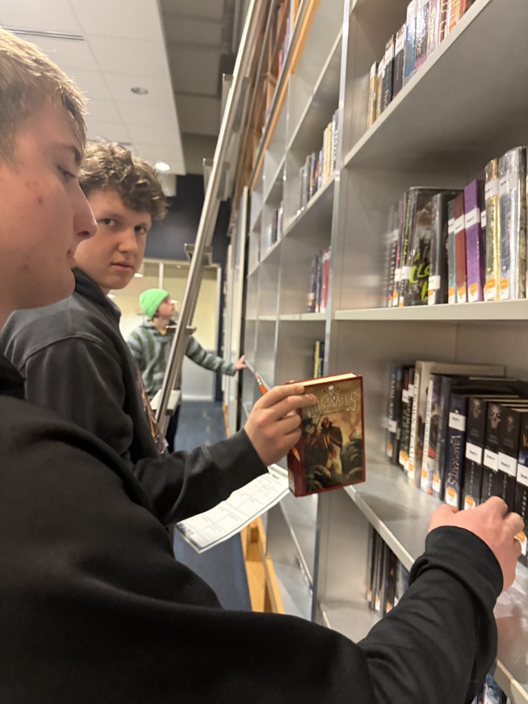 Two boys take books off of library shelves while completing a library scavenger hunt.
