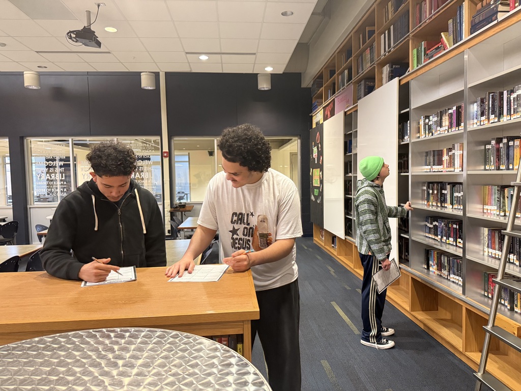 Two boys use a bookshelf as a desk while completing a scavenger hunt in a high school library.