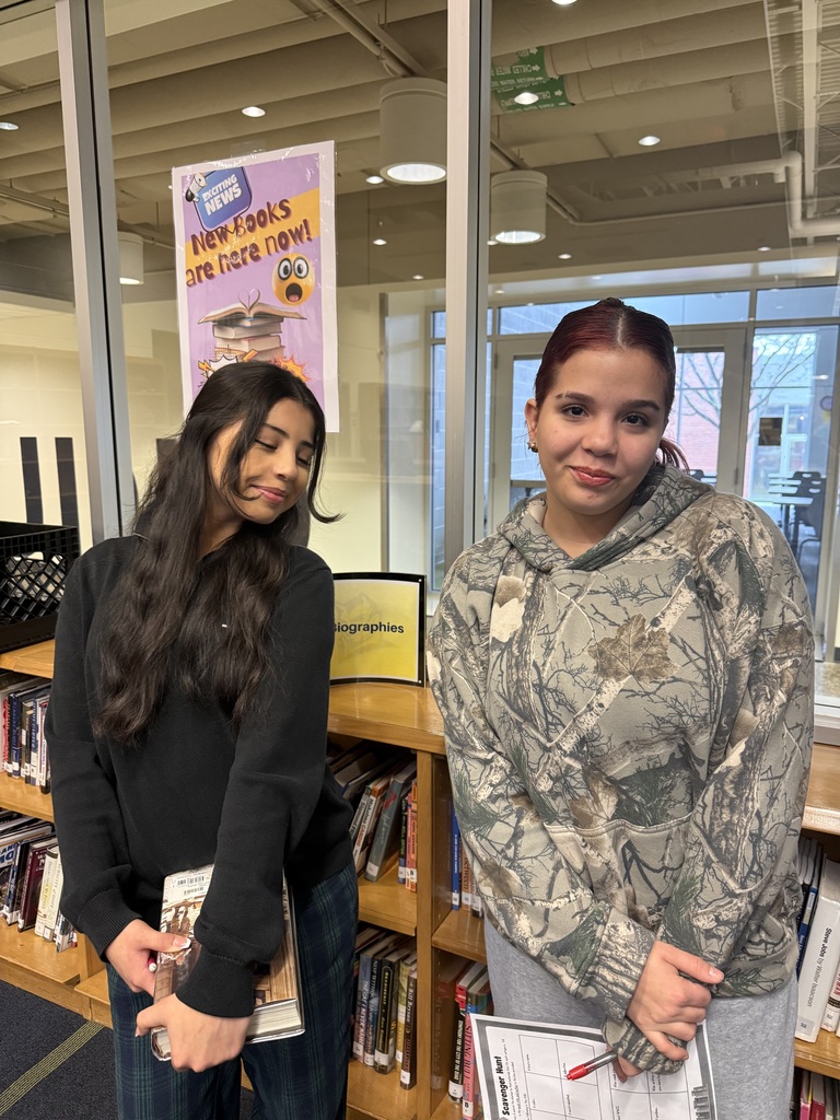 Two girls pose with worksheets in a high school library.