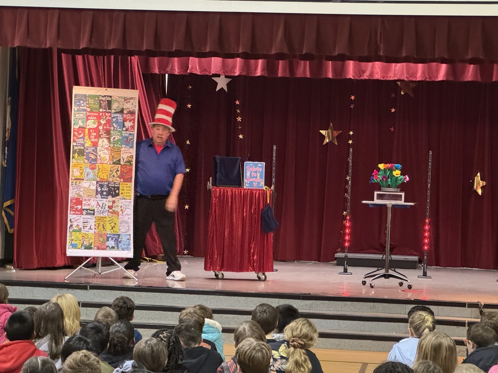A magician wearing a red-and-white striped hat performs on a stage decorated with red curtains and stars during a “Salute to Seuss” magic show for students seated in front of the stage.